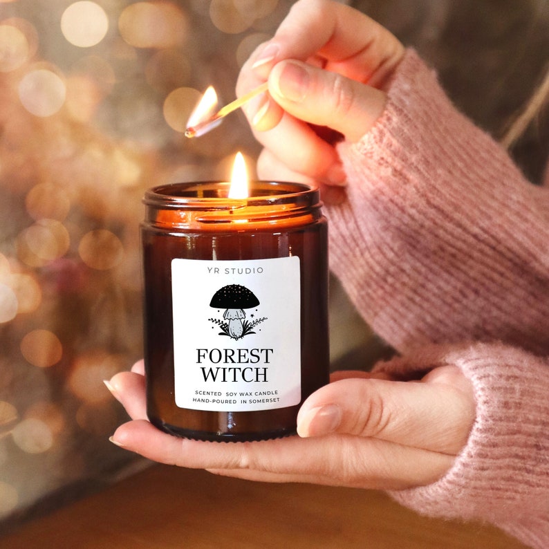 May include: A lit match ignites a soy wax candle in a brown glass jar. The candle has a white label with a mushroom illustration and the words "FOREST WITCH". The candle is hand-poured in Somerset.