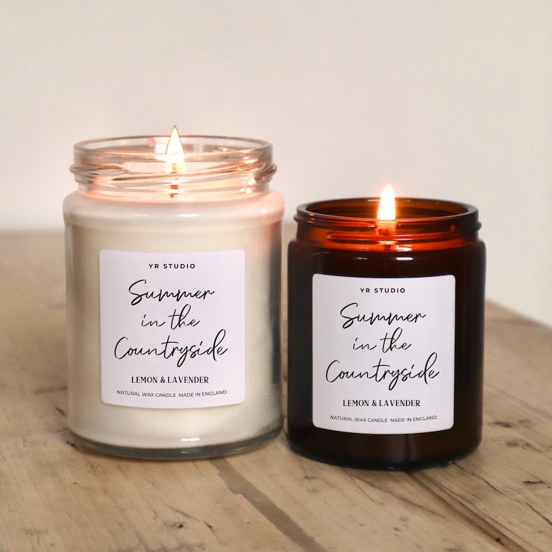 May include: Two candles in glass jars with labels that read "Summer in the Countryside" and "Lemon & Lavender". The candles are lit and the jars are on a wooden surface.