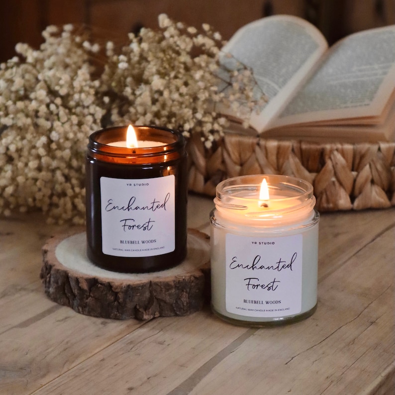 Puede incluir: Dos tarros de velas de vidrio marr&oacute;n con etiquetas blancas. Las etiquetas dicen "Enchanted Forest" y "Bluebell Woods". Las velas est&aacute;n encendidas y las llamas son visibles.