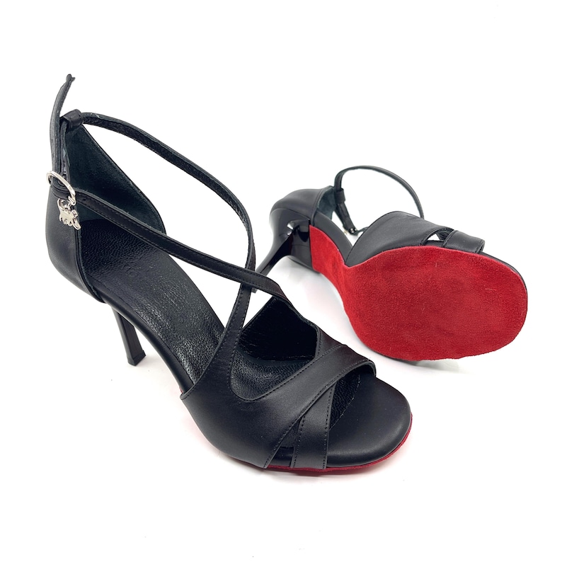 Op de afbeelding: Zwarte leren dansschoenen met een kruisbandontwerp en een dun enkelbandje. De schoenen hebben een rode zool en een kleine hak. De schoenen zijn ontworpen voor ballroom- of latin-dansen.