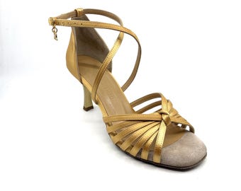 Op de afbeelding: Gouden satijnen dansschoenen met een enkelbandje en een kleine bedel. Het open teenontwerp heeft een beige suède neus en een dunne hak. De schoenen hebben meerdere bandjes over de voet.