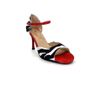 Könnte beinhalten: Schwarz-weiße Zebramuster-Sandalen mit Absatz, rotem Knöchelriemen und roter Wildlederspitze. Die Sandalen haben einen kleinen Metallanhänger am Riemen.