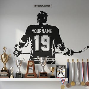May include: A black wall decal of a hockey player wearing a jersey with the text YOURNAME and number 19, positioned above a white shelf displaying various gold and silver trophies, awards, and medals, with a sign reading MY HOCKEY JOURNEY.
