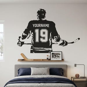 May include: A black and white wall decal of a hockey player in a jersey with the name "YOURNAME" and the number "19". The decal is above a bed with a gray comforter and a wooden shelf with a hockey stick and a framed photo.