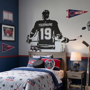 May include: A hockey-themed bedroom with a bed featuring a comforter and pillows with hockey puck and stick designs. A large black hockey player silhouette is on the wall, along with hockey-related wall decor, a hockey stick, and a rug.
