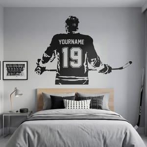 May include: A black and white wall decal of a hockey player in a jersey with the number 19 and the name "YOURNAME". The decal is above a bed with gray bedding and decorative pillows. A hockey stick leans against the wall.
