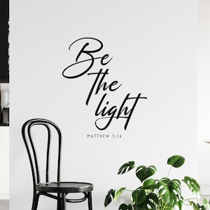 May include: Black and white wall decal with the text "Be the light" and the scripture reference "Matthew 5:14". The decal is displayed on a white wall in a modern kitchen setting.