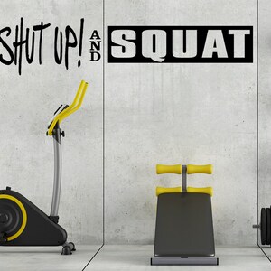 Può includere: Una cyclette nera e gialla con una panca nera e pesi sul muro. Il muro ha una citazione motivazionale che dice "Shut Up! And Squat".