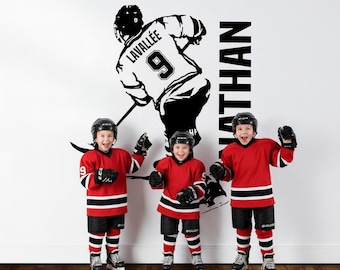 May include: Three children in red and black hockey uniforms and helmets stand in front of a wall decal of a hockey player. The decal includes the name "Lavalée" and the number "9". The name "Nathan" is printed vertically.