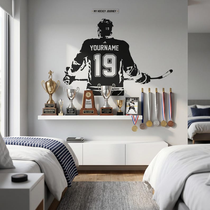 May include: A black wall decal of a hockey player wearing a jersey with the text YOURNAME and number 19, positioned above a white shelf displaying various gold and silver trophies, awards, and medals, with a sign reading MY HOCKEY JOURNEY.