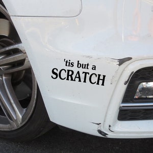 Op de afbeelding: Een witte auto met een zwarte bumper sticker met de tekst 'tis but a SCRATCH'. De bumper heeft wat krassen en slijtage.