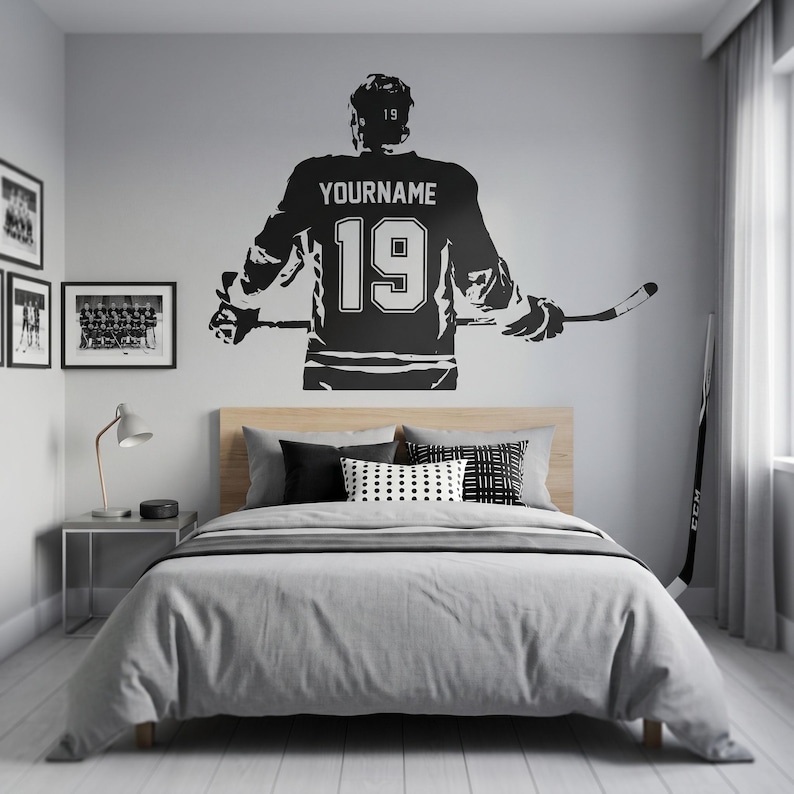 May include: A black and white wall decal of a hockey player in a jersey with the number 19 and the name "YOURNAME". The decal is above a bed with gray bedding and decorative pillows. A hockey stick leans against the wall.