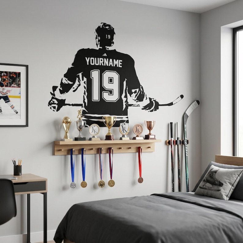 May include: A black hockey jersey wall decal with the name "YOURNAME" and the number "19". A wooden shelf displays trophies and medals. Hockey sticks lean against the wall, and a framed picture of a hockey player is visible.