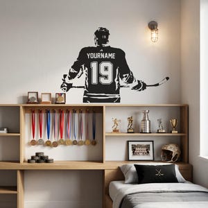 May include: A black wall decal of a hockey player seen from behind, wearing a jersey with the text YOURNAME and the number 19, holding a hockey stick. The decal is positioned above a wooden shelf displaying various sports medals and trophies.
