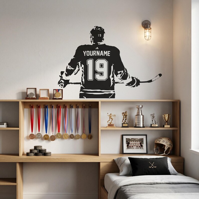 May include: A black wall decal of a hockey player seen from behind, wearing a jersey with the text YOURNAME and the number 19, holding a hockey stick. The decal is positioned above a wooden shelf displaying various sports medals and trophies.