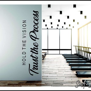 May include: A white wall with black text that reads "Hold the Vision Trust the Process". The wall is in a gym with treadmills in the background.