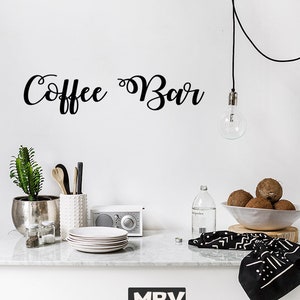 May include: A black wall decal with the cursive text Coffee Bar displayed above a counter. The counter features a silver pot with a green plant, a white textured utensil holder, a stack of white plates, a radio, a bottle, and a bowl of coconuts.