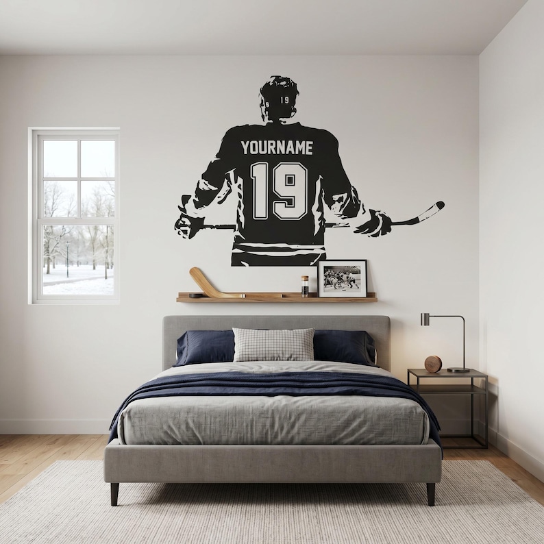 May include: A black and white wall decal of a hockey player in a jersey with the name "YOURNAME" and the number "19". The decal is above a bed with a gray comforter and a wooden shelf with a hockey stick and a framed photo.