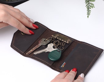 May include: A brown leather key holder with a key ring and multiple keys, a card slot, and a green key fob. The key holder is open and laid flat on a white surface.