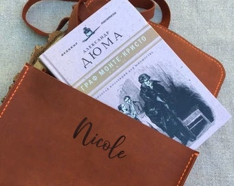 May include: A brown leather book sleeve with the name "Nicole" engraved on the front. The sleeve is holding a book titled "Граф Монте-Кристо" by Александр Дюма.