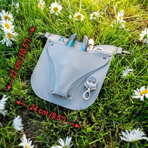 May include: Gray leather tool belt with a 10.5 inch by 9.5 inch measurement. The belt has a pocket for tools and a metal clip. The belt is lying on a bed of green grass and white daisies.