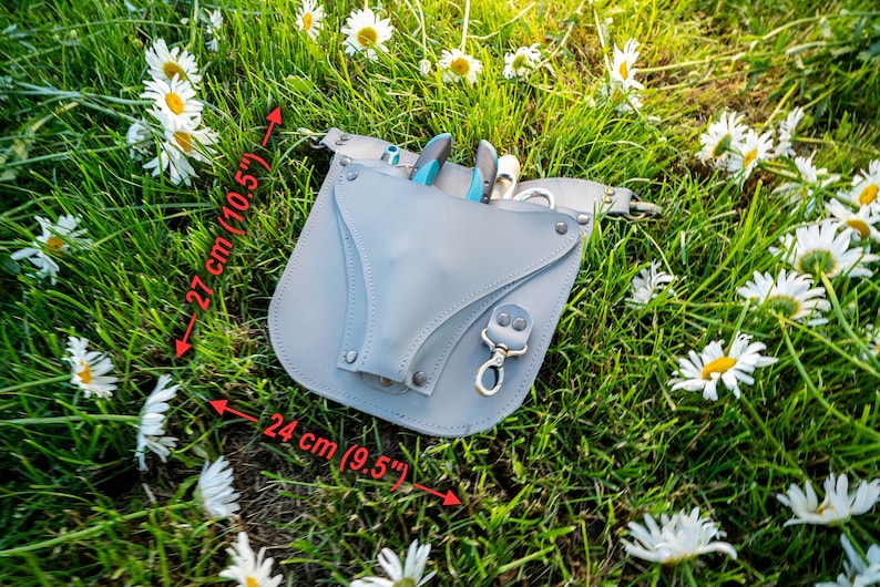 May include: Gray leather tool belt with a 10.5 inch by 9.5 inch measurement. The belt has a pocket for tools and a metal clip. The belt is lying on a bed of green grass and white daisies.