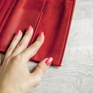 K&ouml;nnte beinhalten: Eine rote Ledergeldb&ouml;rse mit einer Hand, die darauf liegt. Die Hand hat rosafarbenen Nagellack.