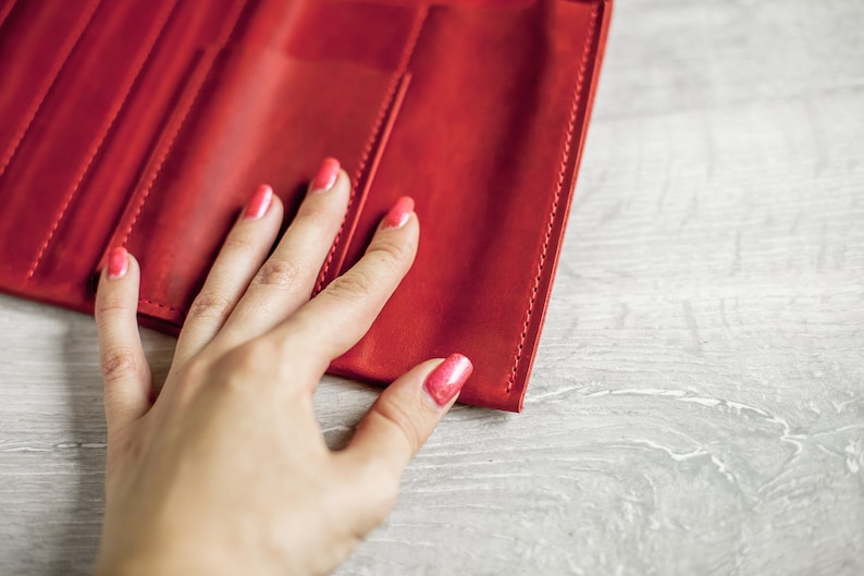 K&ouml;nnte beinhalten: Eine rote Ledergeldb&ouml;rse mit einer Hand, die darauf liegt. Die Hand hat rosafarbenen Nagellack.