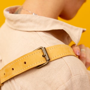 May include: Close-up of a tan-colored jacket with a light yellow leather belt. The belt has a metal buckle and is stitched along the edges. The jacket is made of a soft, textured fabric.