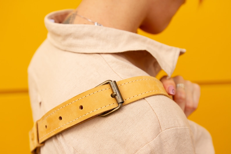 May include: Close-up of a tan-colored jacket with a light yellow leather belt. The belt has a metal buckle and is stitched along the edges. The jacket is made of a soft, textured fabric.