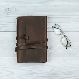 May include: A brown leather journal with a wrap-around tie closure and a pair of silver-framed eyeglasses with clear lenses. The journal is rectangular and lies on a light wood-grain surface.
