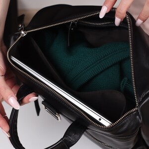 May include: A black leather travel bag with a zippered opening, revealing a laptop and a green sweater inside.