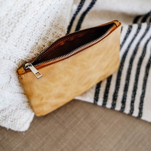 May include: A tan leather zippered pouch with a silver zipper pull. The pouch is open and lying on a white and black striped blanket.