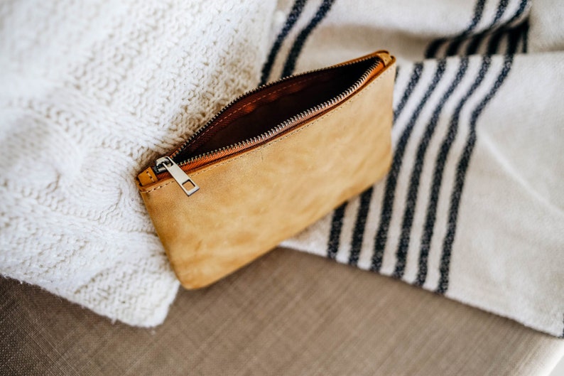 May include: A tan leather zippered pouch with a silver zipper pull. The pouch is open and lying on a white and black striped blanket.