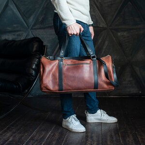以下が含まれることがあります： 黒いアクセントとショルダーストラップが付いた、茶色の革製ダッフルバッグ。 バッグにはジッパー開閉があり、青いジーンズと白いスニーカーを履いた人が持っています。 バッグは新品のようです。