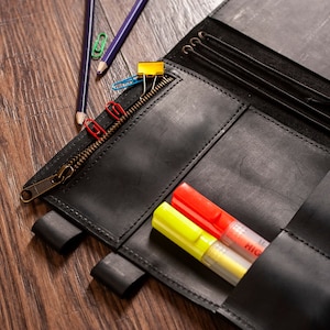 May include: A black leather notebook with a zipper closure and multiple pockets. The notebook is open and has two yellow and red highlighters inside.