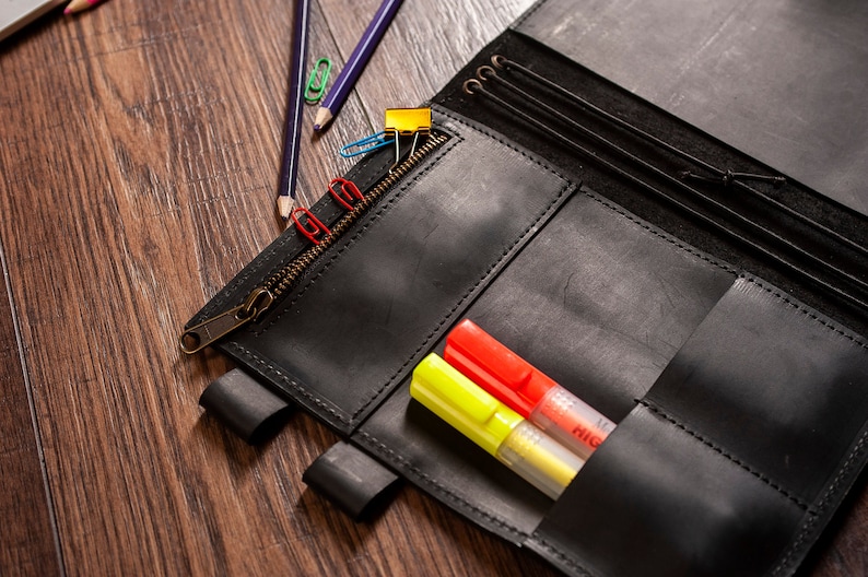 May include: A black leather notebook with a zipper closure and multiple pockets. The notebook is open and has two yellow and red highlighters inside.