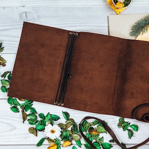 May include: A brown leather journal lies open, revealing its interior. Black elastic bands secure the pages. The journal is surrounded by dried leaves and a white flower. A brown leather strap is visible.