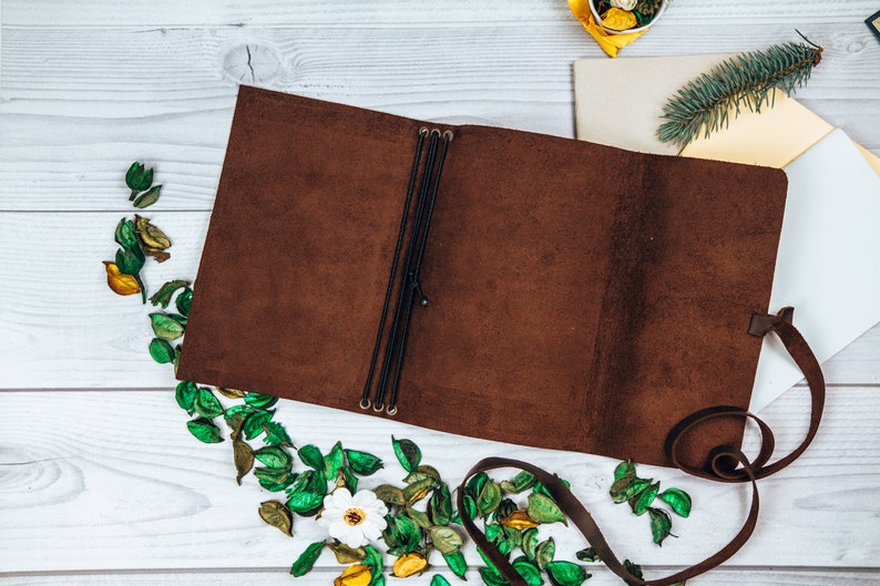 May include: A brown leather journal lies open, revealing its interior. Black elastic bands secure the pages. The journal is surrounded by dried leaves and a white flower. A brown leather strap is visible.