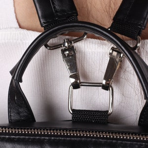 May include: Close-up of the black leather straps and silver buckles of a backpack. The straps are adjustable and have a padded design for comfort.