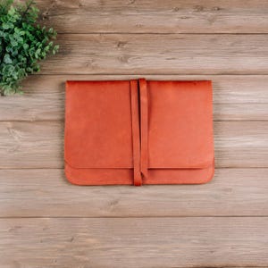 May include: A rectangular, rust-colored leather portfolio or document case featuring a smooth, matte finish and a wrap-around leather tie closure. The item has rounded corners and visible stitching along the edges, presenting a clean, flat design.