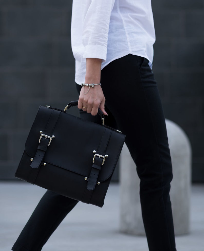 May include: A black leather satchel with silver buckles and metal accents. The bag has a flap closure and a top handle.