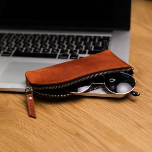May include: A brown leather zippered case with a black zipper and the word "PIKKORE" embossed on the side. Sunglasses are inside the case.