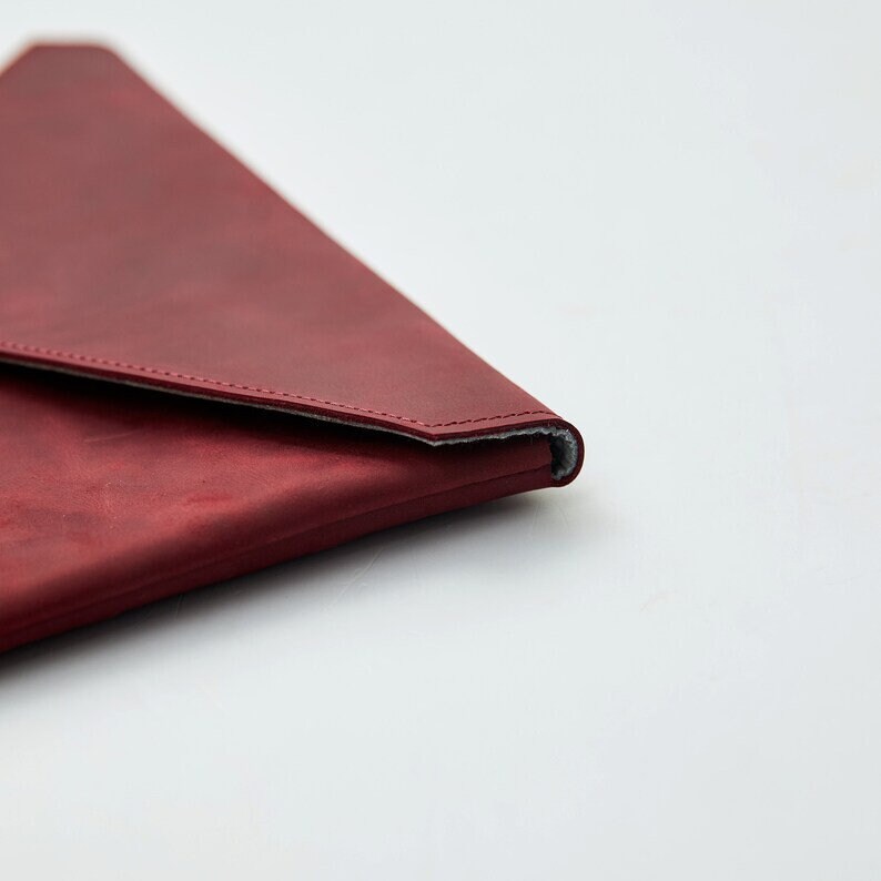 May include: A burgundy leather envelope clutch with a subtle gray lining. The clutch is folded in half and the flap is closed.