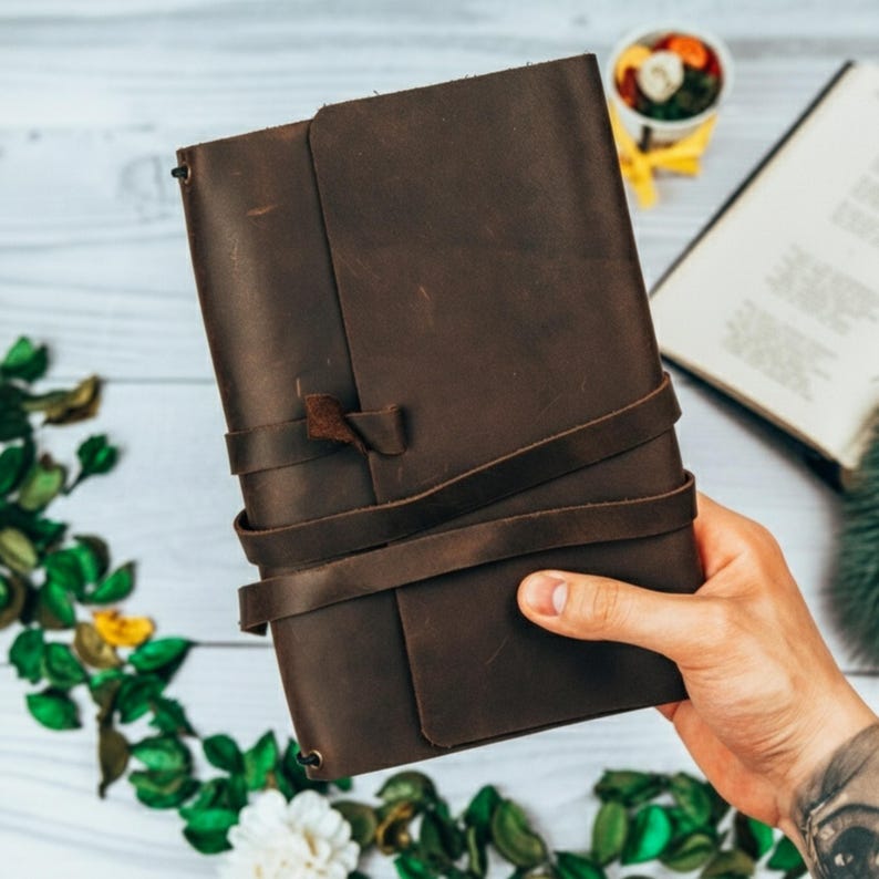 May include: A dark brown leather journal with a wrap-around strap closure. The journal is rectangular and appears to be handmade. The cover has a textured appearance. The journal is held in a person's hand.