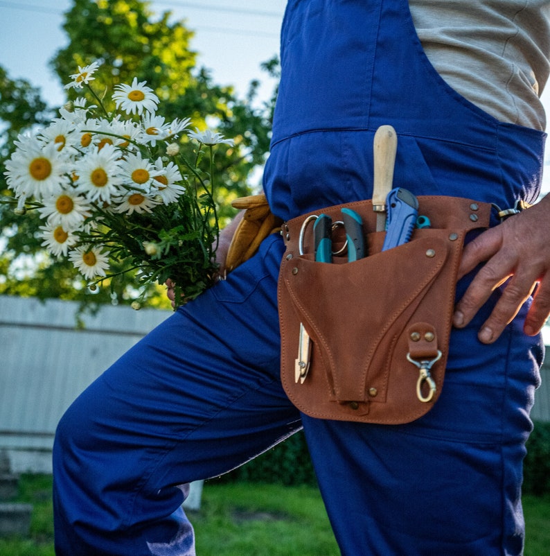 May include: A person wearing blue overalls and a brown leather tool belt with a variety of gardening tools. The person is holding a bouquet of white daisies.
