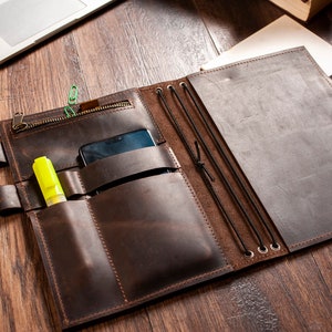 K&ouml;nnte beinhalten: Ein braunes Ledernotizbuch mit mehreren Taschen und Gummib&auml;ndern. Das Notizbuch hat eine Rei&szlig;verschlusstasche auf der Vorderseite und eine Telefontasche im Inneren. Ein gelber Textmarker und ein schwarzes Telefon sind in den Taschen sichtbar.