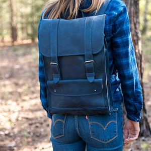 May include: A dark blue leather backpack with two adjustable straps and a front pocket. The backpack is worn by a person in a forest setting.