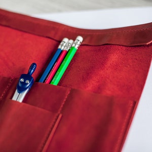 K&ouml;nnte beinhalten: Ein rotes Leder-Bleistift-Etui mit vier Bleistiften und einem Zirkel. Die Bleistifte sind blau, rot, gr&uuml;n und wei&szlig;.