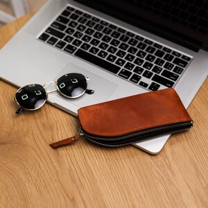 May include: A brown leather zippered case for glasses with the word "DIVORE" embossed on the front. The case is sitting on a wooden surface next to a laptop and sunglasses.
